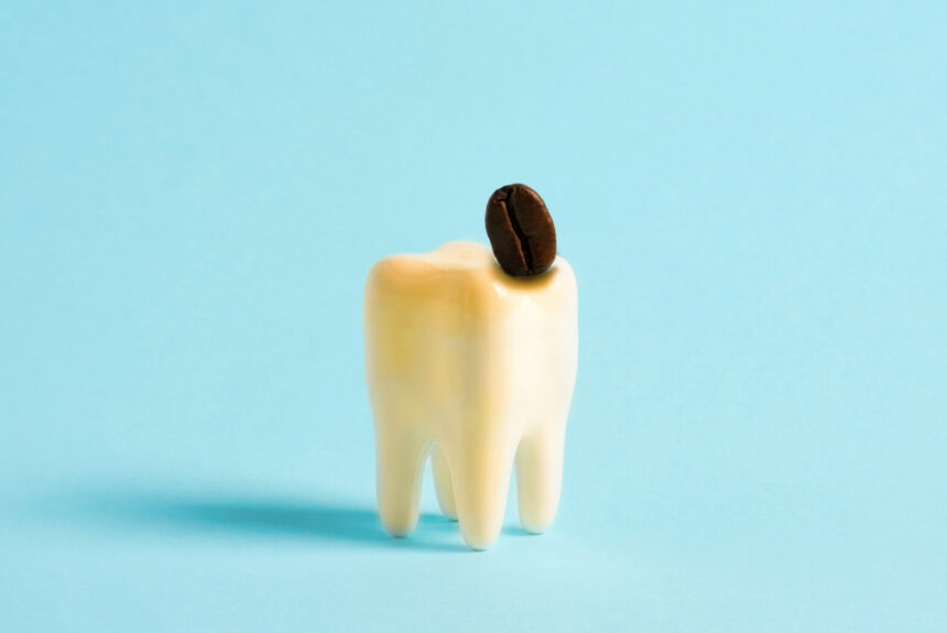 A model of a human tooth with yellow discoloration and a single coffee bean on top, set against a blue background.
