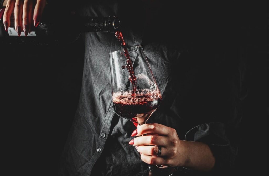 An individual pouring red wine into a glass against a dark background, illustrating a habit that can stain teeth.