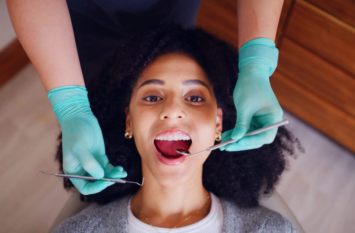 An overhead view of a woman with curly hair smiling in a dental chair while a dentist in teal gloves performs a recall exam with a mirror and probe.