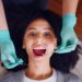 An overhead view of a woman with curly hair smiling in a dental chair while a dentist in teal gloves performs a recall exam with a mirror and probe.