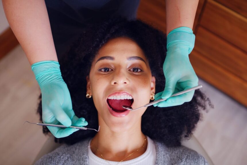 An overhead view of a woman with curly hair smiling in a dental chair while a dentist in teal gloves performs a recall exam with a mirror and probe.