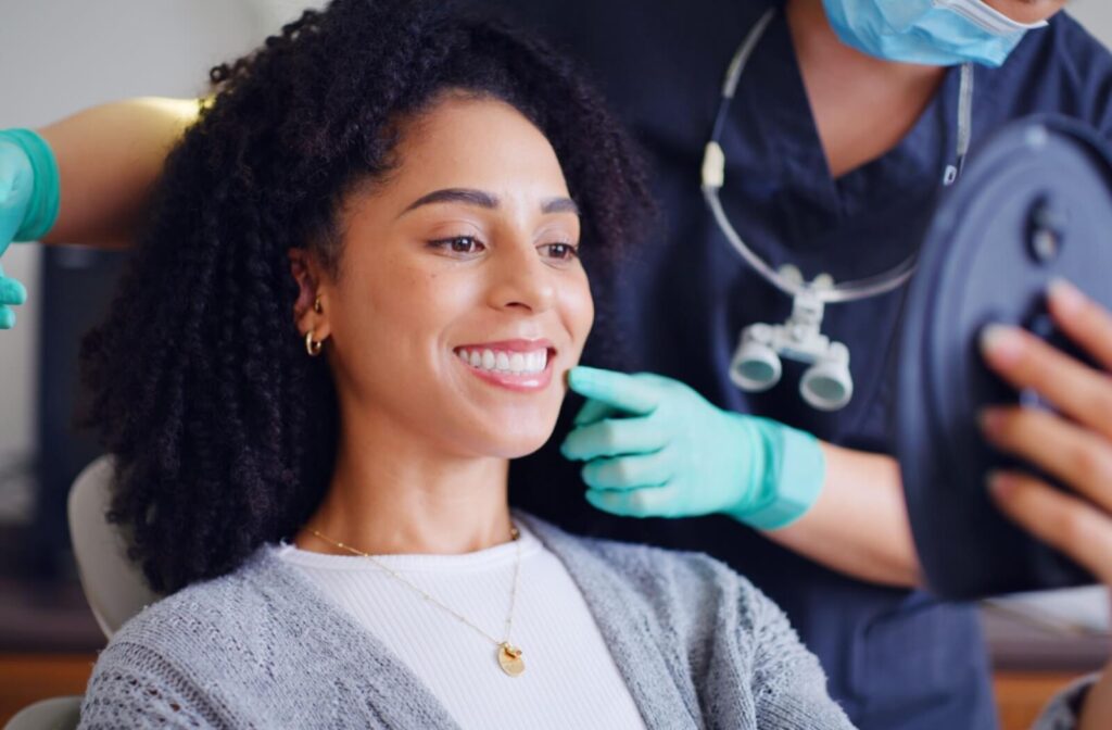 A smiling woman with curly hair looking in a handheld mirror as her dentist points to her healthy teeth after a dental exam.